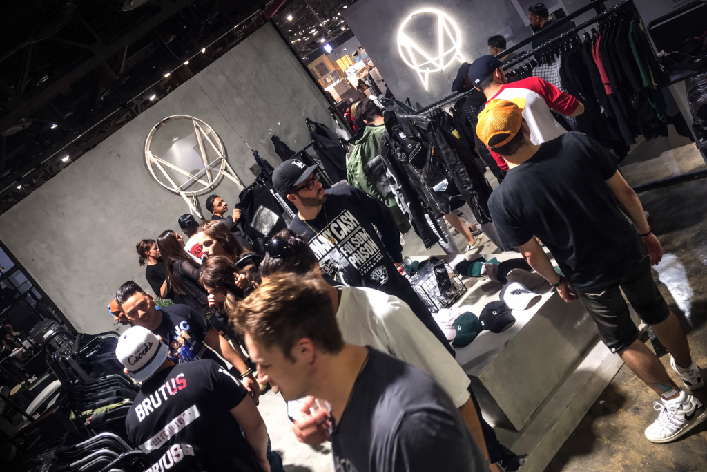 Crowd browsing streetwear inside ComplexCon retail activation with illuminated neon M logo on concrete wall